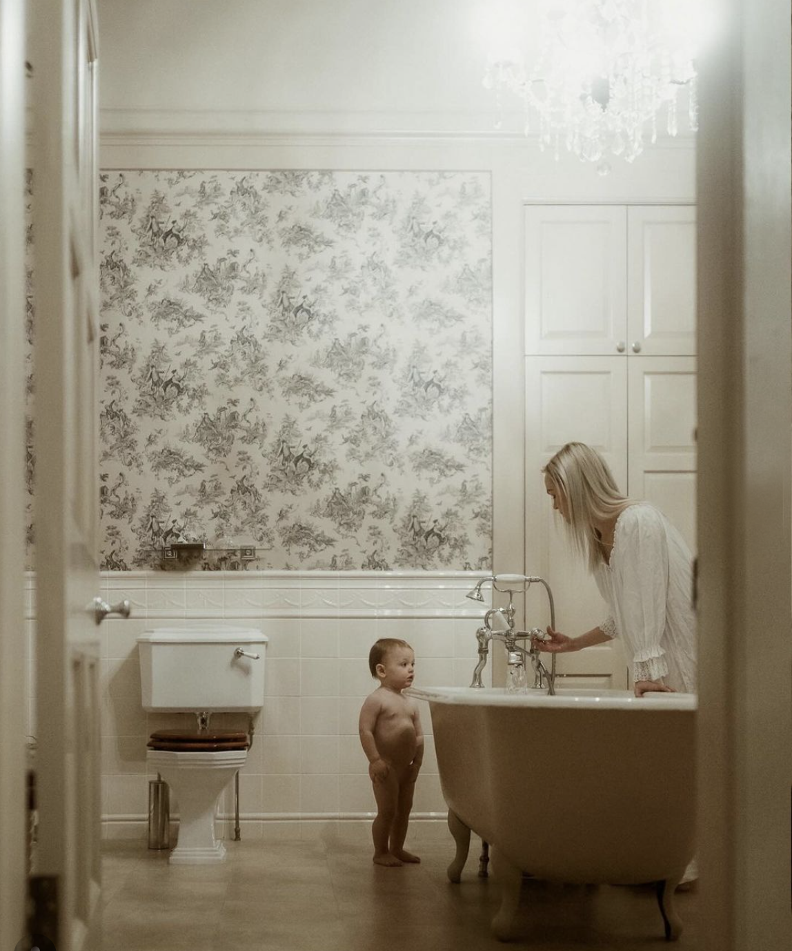 Mother and baby toddler in bathroom with clawfoot bathtub and floral wallpaper